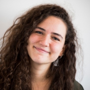 A woman with long, curly brown hair smiles softly with her head slightly tilted. She wears small hoop earrings and a dark green shirt. Plain light grey background.